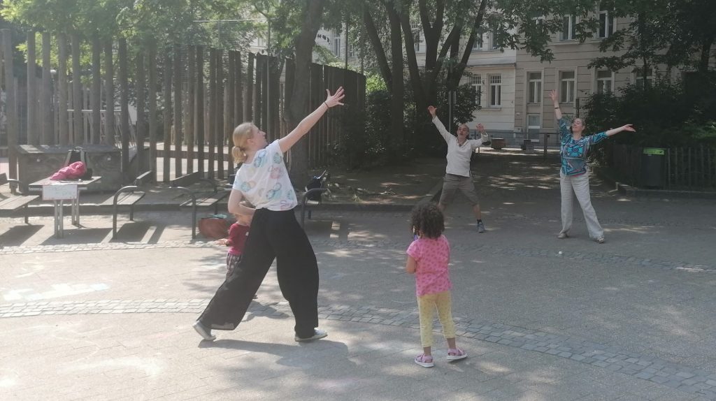 Das Bild steigt tanzende Menschen mit nach oben seitlich weit ausgestreckten Armen in eienm Park. auch Kinder sind dabei.
