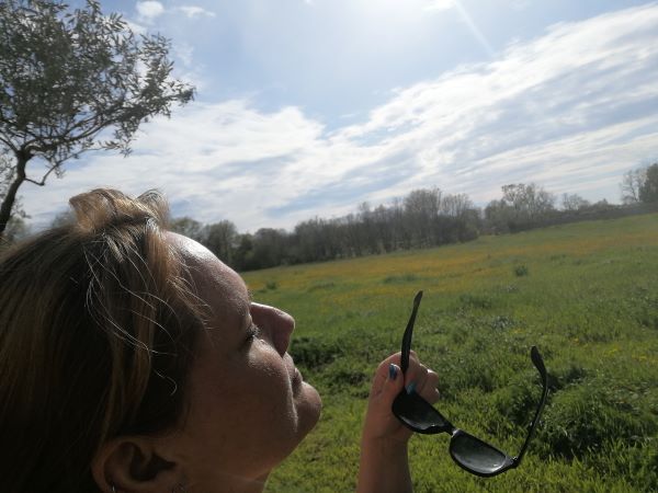 Auf dem Bild ist eine Wiese in der Sonne zu sehen mit Bäumen am Rand und blauem Himmel mit ein paar weißen Wolken. Vorne links ist Lorenas Gesicht der Sonne zugewandt. Sie hält eine Sonnenbrille in der Hand.