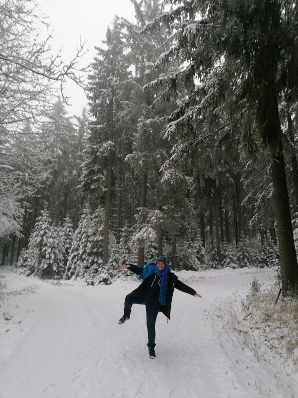Lorena steht in einem winterlichen Wald mit Schnee und hohen Bäumen auf einem Bein.