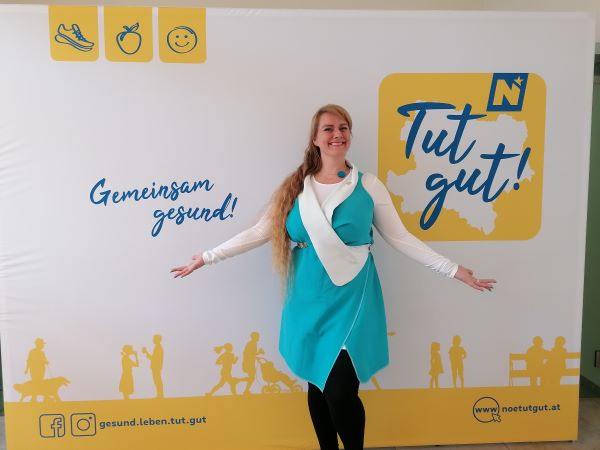 Lorena vor einer Fotowand von "Tut gut!" in einem türkisen Kleid, die Arme ausgebreitet, lachend.