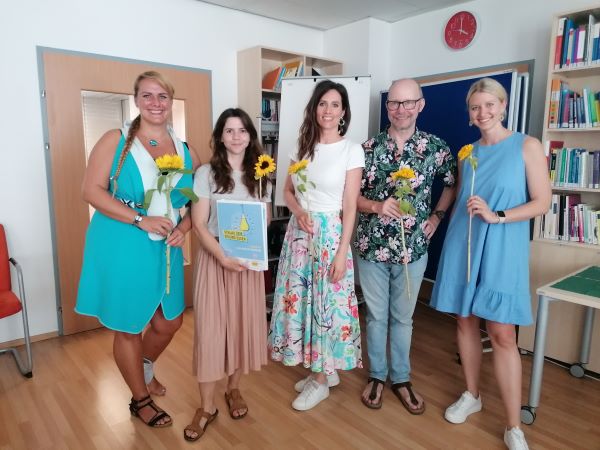 5 Menschen nebeneinander, sie halten Sonnenblumen in den Händen und eine Mappe auf der "Schlau se3in - gesund essen" steht. Lorena ist ganz links zu sehen.